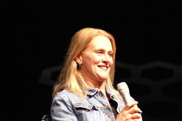 A woman in the spotlight on a stage. It's a close-up of her from the shoulders up. She's looking to the right side of the snap. Smiling, while holding a microphone just below her face. She's got long blonde hair and she's wearing denim jacket.