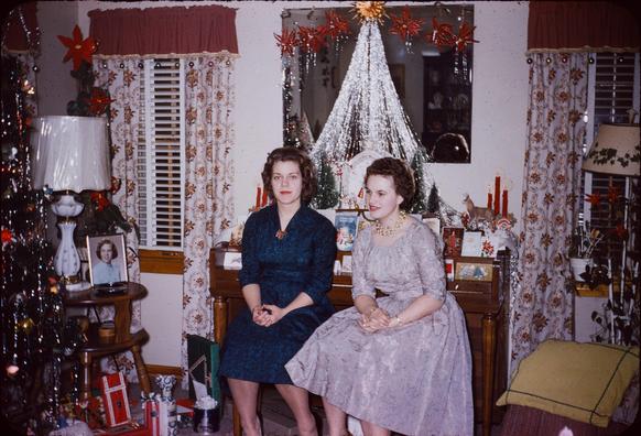 A woman in blue and a woman in lavender sit together on a piano bench in a room with a Christmas tree and numerous other seasonal decorations.