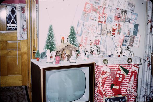 A room decorated for Christmas. The wall by the TV is covered in numerous holiday cards. A small nativity scene is set up on top of the TV. On the TV is a dimly visible image of a man in a dark suit standing near a woman with dark hair in a light colored dress. The image is black and white.