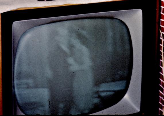 Cropped view of the TV screen with the contrast brought up to make the darker areas darker. Now we can see that the man appears to have male pattern baldness and might be wearing glasses.