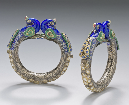 photo of a pair of beweled bangle-style bracelets on grey background, one in front view and one in side view, each topped with a pair of peacocks, one facing right and one left