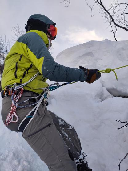 A figure in a yellow jacket wearing reflective ski goggles hangs on a rope over a small cliff. Everything in the background is covered in snow
