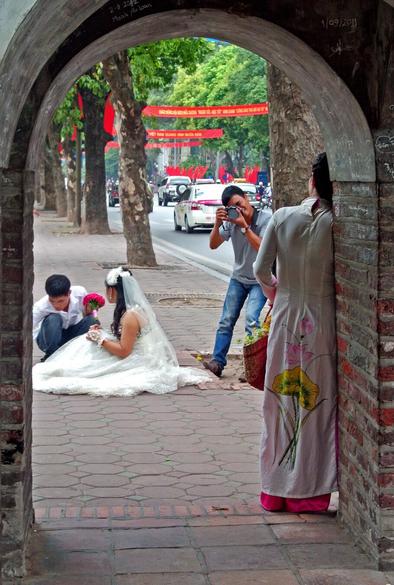 Photo shows a bridal photo session with two brides and two photographers, seen through an old gate in the old four gates tower. One bride is posing in the gate dressed in a classic female Vietnamese Ao Dai decorated with flowers. In the background a bride in white European style wedding dress is sitting and getting styled for a photo shoot. In the background in an alley of trees towards, bordering a three field city road to the right, and a walkway with green bushes to the left. It is a slightly overcast day, but the colours of the bridal clothes gives off a nice contrast.