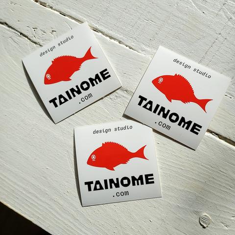 Three vinyl stickers on a wooden table, the stickers present the icon of a bright red fish in the middle and the texts "tainome.com" and "design studio" below and above it, set in two different custom typefaces (one massive and geometric-looking, the other light, monospaced and presenting inktraps).