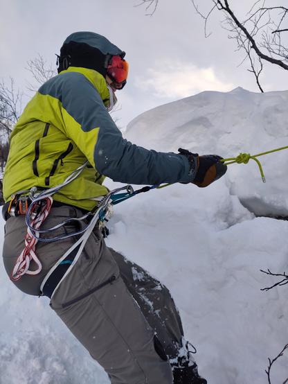 A figure in a yellow jacket wearing reflective ski goggles hangs on a rope over a small cliff. Everything in the background is covered in snow