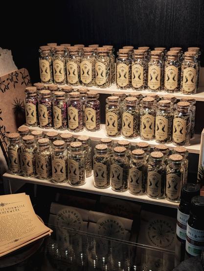 Das Foto zeigt ein kleines Holzregal, das in einer Vitrine steht. Das Regal ist voller kleiner Glasbehälter mit Korken, die mit einer Vielfalt an getrockneten Kräutern und Räucherwerk gefüllt sind.