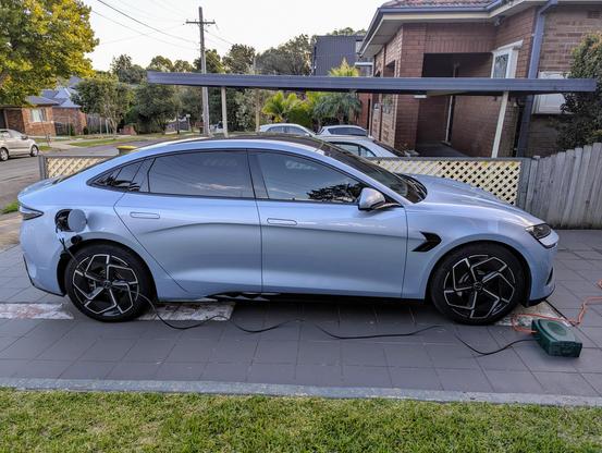 Photo of a BYD Seal electric sedan car parked in a driveway. There is a black cable running from the charging port at the rear of the car to a green box on the ground by the front of the car, and an orange cable emerging from that box and continuing out of shot.