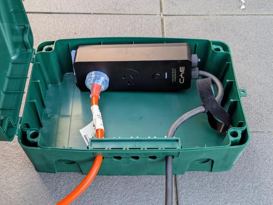 Photo of a green plastic box, open, with a small black BYD branded 2 outlet mains power board inside it. An orange extension cord is plugged into one of the two outlets.