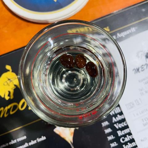A close-up of a shot glass filled with Sambuca with three dark coffee beans floating inside. The glass sits on a restaurant table above a printed menu, suggesting this is the end of a meal. The reflection and clarity of the liquid are captured crisply.