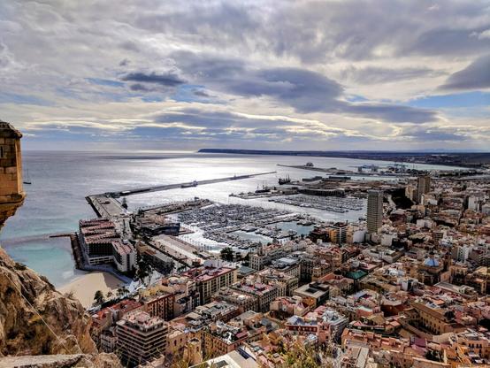 A high-angle panoramic photograph shows a coastal city with a harbour full of boats, viewed from a rocky elevation. The foreground features a portion of a stone structure on the left. Below it is a small, sandy beach with turquoise water. The city sprawls across the middle ground, a dense collection of buildings with terracotta roofs, interspersed with taller modern structures. A large harbour with numerous white boats fills a significant portion of the frame, extending into a calm, grey sea under a cloudy sky. The horizon line is visible in the distance, where the sea meets the sky.