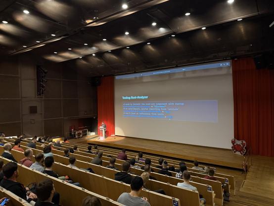 A presentation taking place in a theater-style venue, with a speaker addressing an audience. The screen displays code and information related to "Tooling: Rust Analyzer." The audience appears engaged, and a crab-themed decoration is visible near the stage.