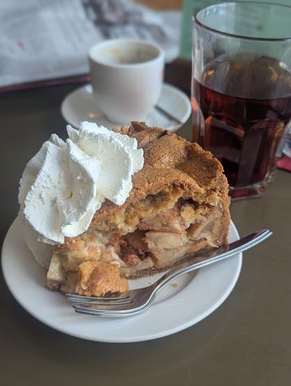 a white plate with a piece of apple pie and a big dollop of cream. two forks lie on the plate as well. behind the pie there is a glass of black tea and a cup of coffee. the table is Light-Brown/olive green.