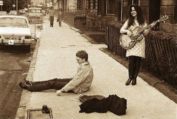 A sepia tinted photo of an urban street. Alan Horne sits on the pavement, his legs out in front of him, his bag and jacket on the floor beside him. Behind him, a smiling Jill Bryson is playing a stylish semi acoustic guiter. She's wearing a light coloured polka dot dress and dark trousers. In the distance, two people are walking towards them.