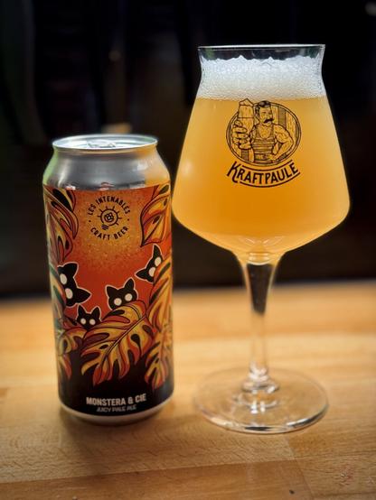 A can of Les Intenables: Monstera & Cie Juicy Pale Ale next to a full beer glass. The beer is indeed pale with a frothy white head. The glass reads Kraftpaule with a cartoon drawing of a burly man holding a beer glass.