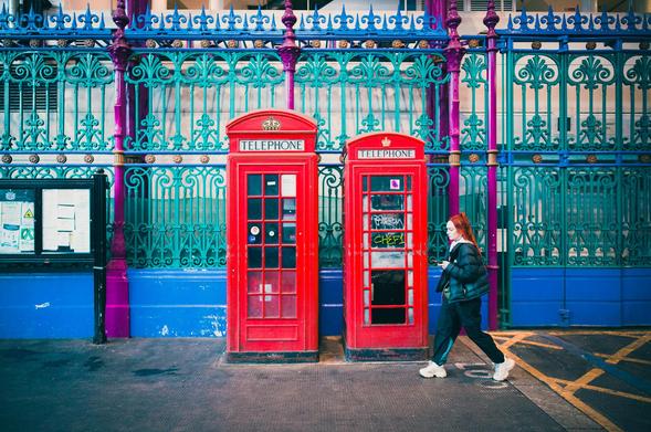 A young woman with red hair is walking past two red telephone booths. A decorative iron gate serves as a backdrop, painted in shades of blue, green, and purple. In the foreground, there is a notice board and painted yellow lines on the ground.