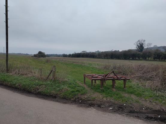 Run of trees in the distance that show the line of the former railway approaching the bridge site over the road in the forground.