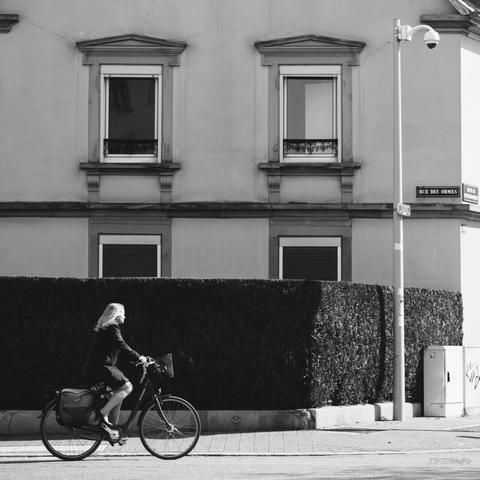 A squared black and white picture of a blond lady on a bike passing in front of a house with a dark contrasty hedge, in Rue des Ormes, Strasbourg. In the opposite corner of the picture, a pole with a camera is monitoring the street.