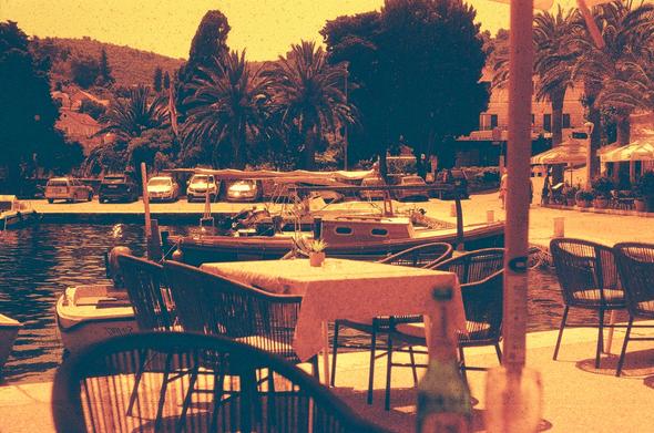 A redscale photo taken from outdoor bistro seating, the foreground contains a couple of empty seats; the midground has several parked boats and then the background has several parked cars and trees obscuring the rest of the view, leaving a few homes and a foresty hillside visible
