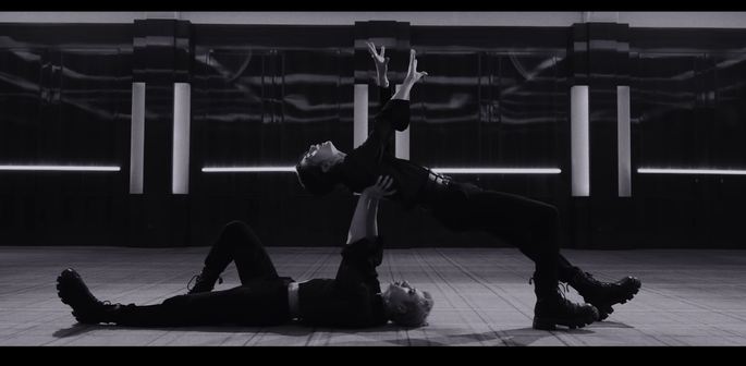Bang Chan lying flat on his back, supporting Hyunjin in a dance move where he is leaning back and resting all his weight on Chan, while reaching towards the ceiling himself