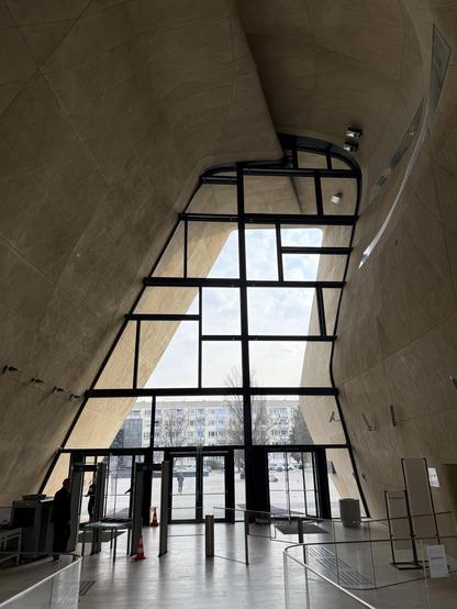 Interior of a modern building featuring curved walls, large floor-to-ceiling windows, and a security check area. Natural light filters in, illuminating the space. Outside, a street view with buildings and a tree is visible through the glass.