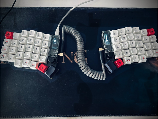A split keyboard called a soflev2 is placed on a large mouse mat, a long cable connects the two halves, coiled, and the keycaps are mostly white with some black and red as well.