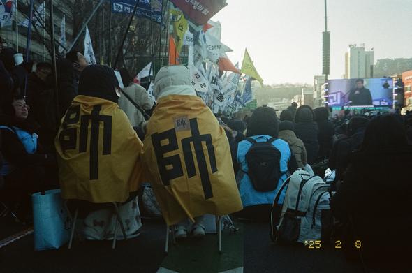 2025년 2월 8일 광화문 광장. '수자기'의 수(帥) 글씨를 망토처럼 두른 두 시민이 간이의자에 앉아있다. 앞에는 많은 사람들이 앉아 시민발언을 듣고 있고, 왼편에는 각종 깃발들이 휘날리고 있다.