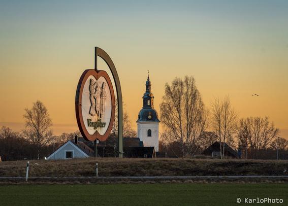 A sign shaped like an apple, welcoming people to the town of Vingåker. A church in the background.
