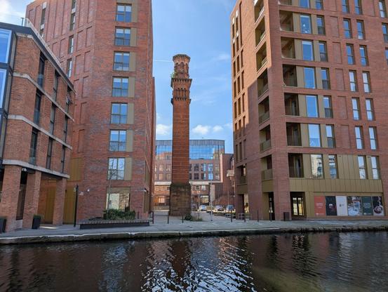 In-between two modern brown brick apartment buildings stands a beautiful old Venetian style tower standing tall. In front of all is the tow path and canal.