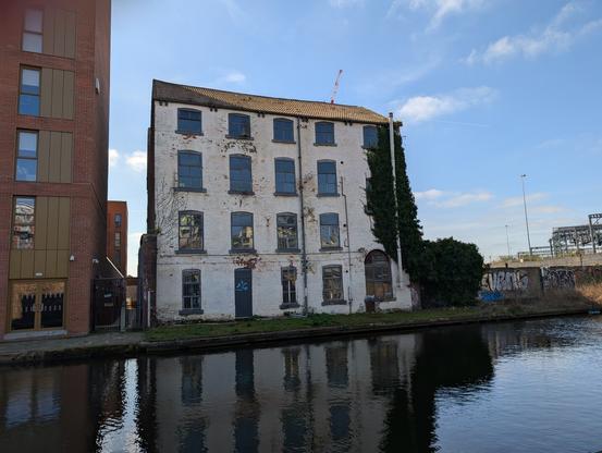 On the other side of the modern brown apartment buildings is an older dilapidated Victorian building with ivy growing up it.in front is the canal and behind blue sky with wispy white clouds