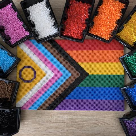 The Progress Pride flag made out of Hama beads. The flag has rainbow stripes on the right side and white, pink, bright blue, brown and black stripes on the left and middle. On the very left there is a purple circle on yellow. The flag is surrounded by little boxes full of beads in the colors I used.