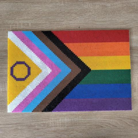 The Progress Pride flag made out of Hama beads. The flag has rainbow stripes on the right side and white, pink, bright blue, brown and black stripes on the left and middle. On the very left there is a purple circle on yellow.