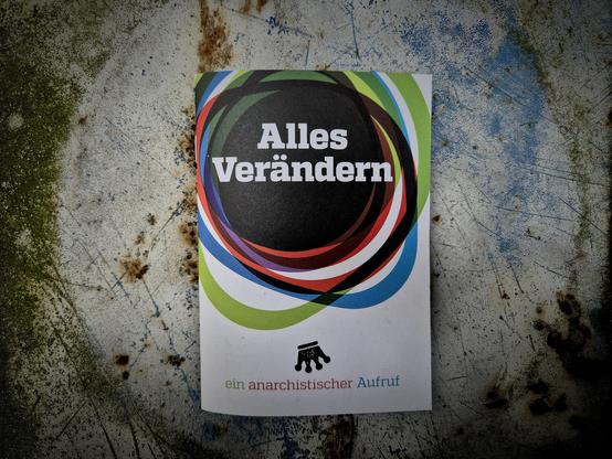 Eine anarchistische Broschüre mit dem Titel „Alles Verändern – ein anarchistischer Aufruf“ liegt auf einer verwitterten, rostigen Metallfläche. Der Titel ist von bunten, sich überschneidenden Kreisen eingerahmt.

English:
An anarchist brochure titled “Change Everything – an Anarchist Call” lies on a weathered, rusty metal surface. The title is framed by overlapping, colorful circular shapes.
