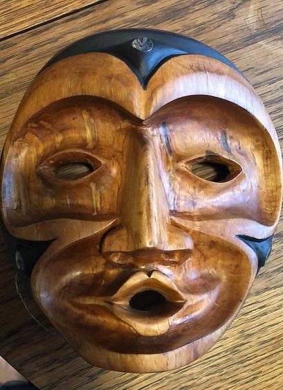 Close-up of a Indigenous Coast Salish handcrafted, expressive wooden mask resting on a wood surface. The mask has a rounded face, stylized features with eye-holes and an open mouth. Black painted sections accent the edges, and a round piece of abalone shell is embedded at the center top. The wood grain is clearly visible, suggesting a natural finish.