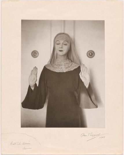A scan of a cabinet card photo of a woman wearing something like a secular nun's outfit (sorry, I'm bad with fashion terms) with her hands raised to shoulder height, palms out.