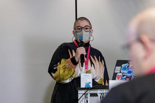Anna e só, a young white femme person in their early 30s, holds the microphone in front of the stage near a stand with their laptop full of stickers as they talk about Outreachy. They have very short hair they're wearing prescription glasses with black frames with a grey, valved N95 mask. They're wearing a black shirt with a beautiful illustration of big Mandacaru flowers in their chest layered with a black turtleneck and a black long skirt. The pink lanyard of the conference creates a nice contrast against their clothes. In the foreground, but blurred, Jarek (the moderator of the stage—an older white man in a black t-shirt) watches Anna speak.