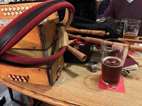 A pub table with an old Hohner pressedwood diatonic accordion and a box of bagpipes on it, along with a couple of beers. You might make out a pair of Anglo concertinas in the background too.