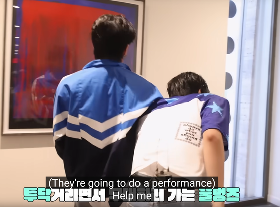 Changbin walking away from the camera while holding Seungmin's shoulder under his arm. Caption: (They're going to do a performance) Help me