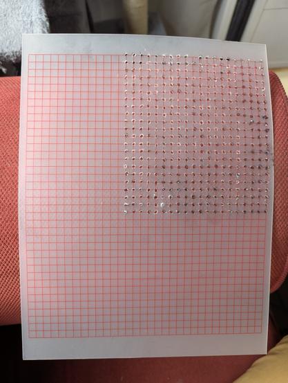 A transparent A4-sized sheet of plastic, with a grid printed on it. Holes have been poked into about a quarter of the sheet on the intersection points of the grid.