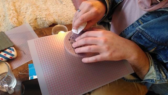 A photo of me poking holes into a sheet of transparent plastic with a grid printed on it. A candle sits next to me, for heating the needle.