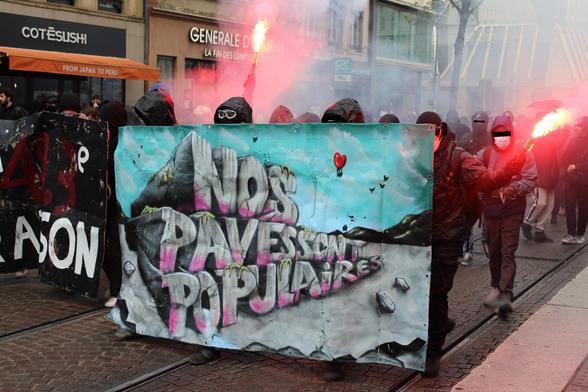 Banderoles "Nos pavés sont populaires" et "Ma 49.3e raison" et des manifestant•es du bloc avec des fumigènes rouges du côté de Alt Winmärik.