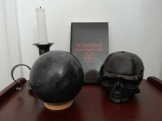 A wooden surface against a white wall, containing a white candle in a blackened metal holder , a black granite ball, a copy of The Satanic Rituals by Anton Szandor LaVey (black cover, lettering and Sigil of Baphomet in red) and a black skull ornament, lower jaw absent.