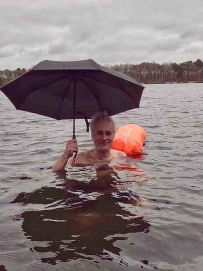 Person mit Regenschirm in der Hand steht im See mit Wasser bis zu den Schultern. Man sieht eine Schwimmboje und den stark bewölkten Himmel.