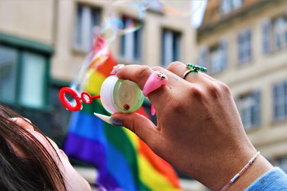 Une personne souffle dans un tube qui fait des bulles. En arrière-plan, un drapeau rainbow