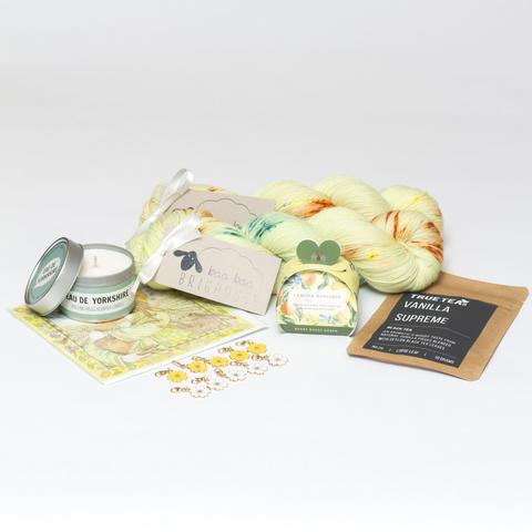 Picture shows two skeins of yarn, one behind the other, in shades of lemon with yellow, orange and green speckles. In front of the yarn are a number of gifts including (from left to right) a tin candle, a card, flower stitch markers, a gift box containing soap and a packet of vanilla tea.