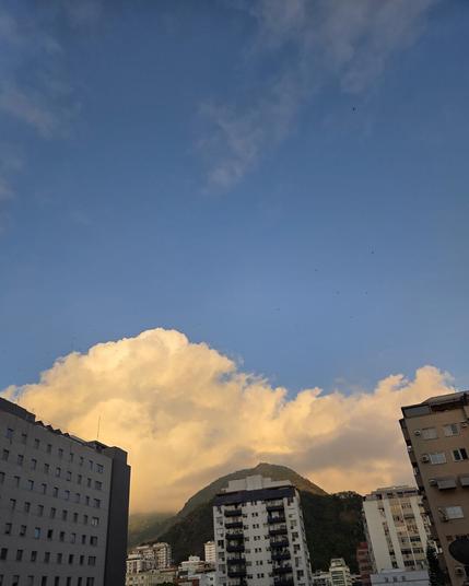 Vista a partir da minha janela. Emoldurando o cenário, vários prédios e o morro do Corcovado ao fundo, quase totalmente coberto por nuvens. O céu azul da manhã contrasta com as nuvens amareladas que encobrem o morro, criando uma atmosfera tranquila e inspiradora.