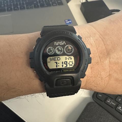 Round digital watch with black resin case cover and bezel, two pushers on each side, digital display in bottom of the black screen showing day of the week, month and day, and hours-minutes-seconds. Three round digital subdials in the top half of the screen: two smaller ones with blue-green background showing state of charge and active functions, the larger one with pinkish-red backgound showing a seconds countdown that repeats every ten seconds. The NASA "worm" logo is at the very top of the screen.