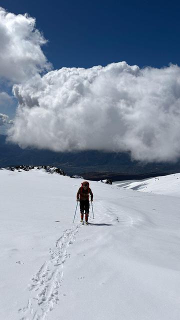 A skier ascends some white snowfields. behind him is a big bank of white cloud