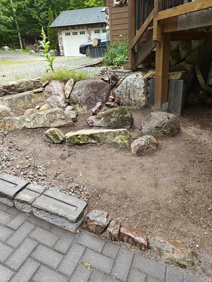 View from below of a small unplanted rock garden on a slope   tucked into the corner of a curving paved path. The large rocks from the local area have spaces between for planting and give way to a shallow sloped scree below.