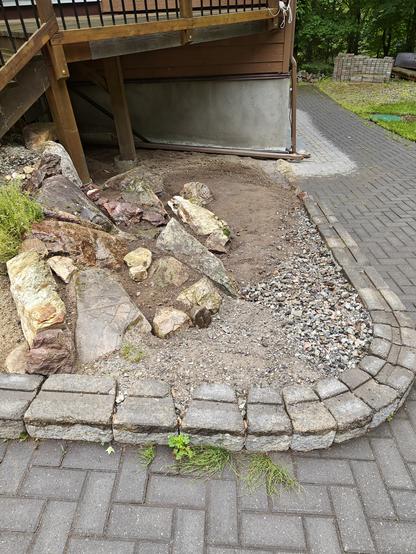 View from above of a small unplanted rock garden on a slope   tucked into the corner of a curving paved path. The large rocks from the local area have spaces between for planting and give way to a shallow sloped scree below.