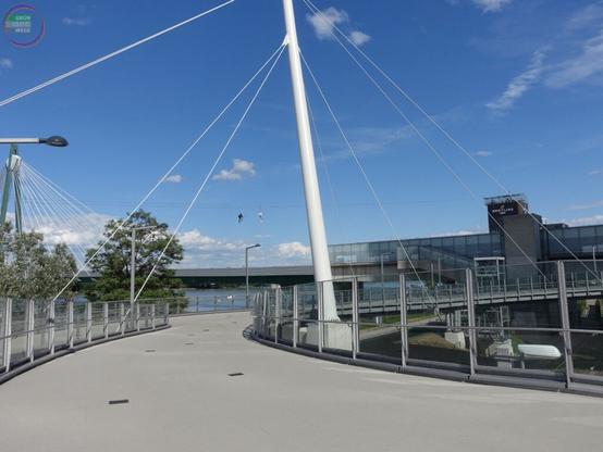 Kurvige Brücke, dahinter ein Fluss, entlang schweben zwei Menschen auf einer Seilrutsche